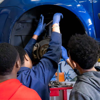 Three young people working together to inspect and repair a brake assembly on a raised blue car in an automotive workshop